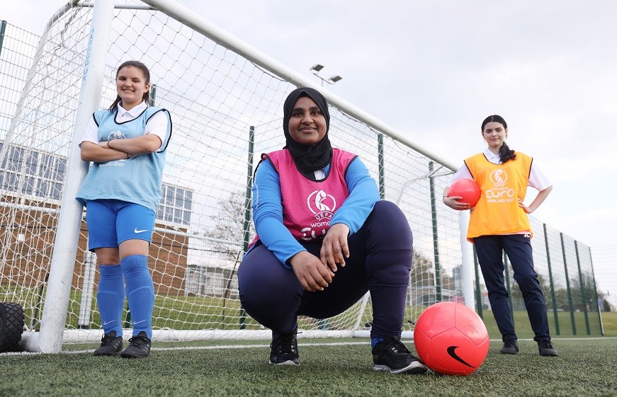 Three girls ready to play football. They are in front of goal posts.