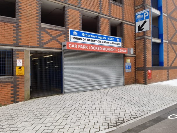 Outside of car park with grey shutter down with signs that read 'Grosvenor Square MSCP Car Park Locked Midnight - 5:30am'