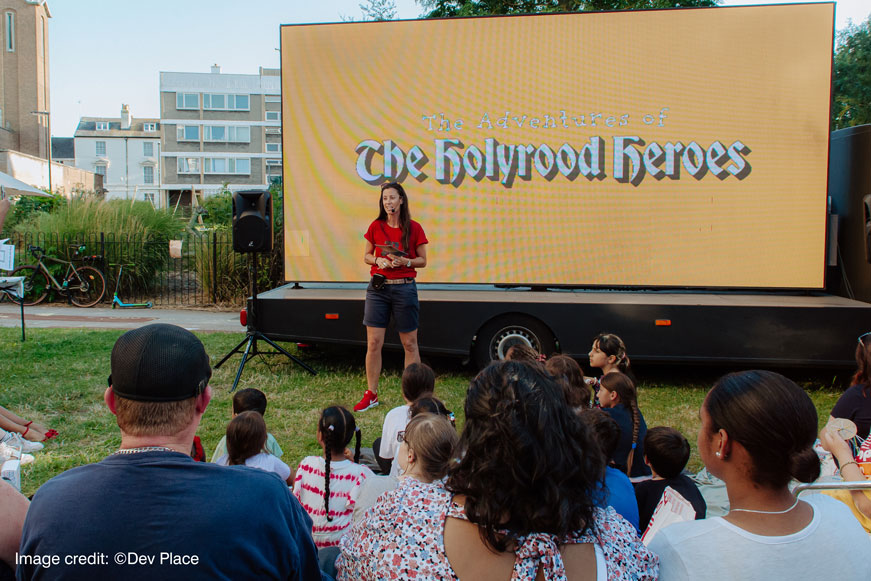 A Crowd Looking At A Woman Standing In Front Of A Screen That Says: The Adventures of The Holyrood Heroes. Image credit: copyright Dev Place