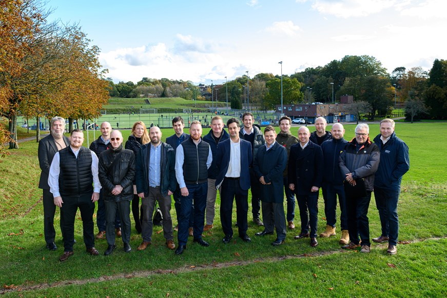 Image of group of men and a woman standing on a green field all smiling. Behind the people are green courts and trees with green and brown autumnal leaves.