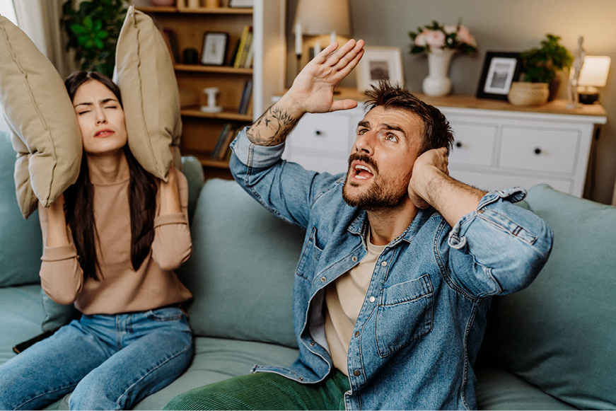 One Person With Cushions Over Their Ears And Another With One Hand Covering An Ear Exclaiming Towards The Ceiling