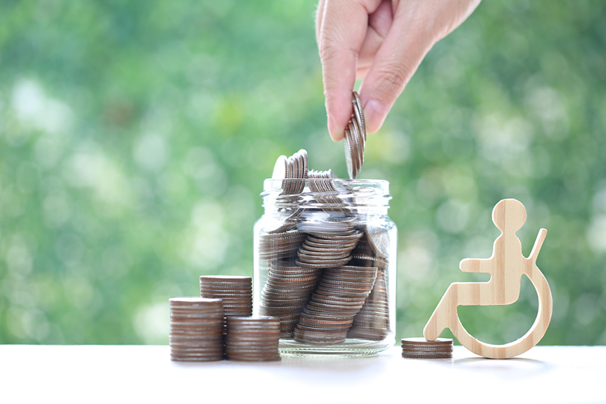 Glass Jar Filled With Money With A Wooden Disability Symbol Next To It Glass Jar Filled With Money With A Wooden Disability Symbol Next To It