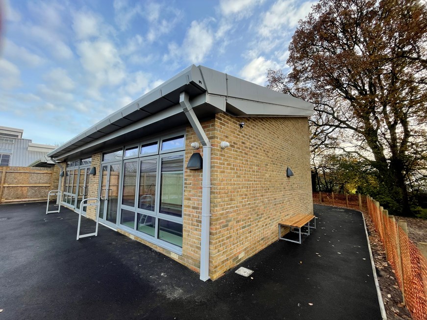 Image of outside of brown brick building with grey windows and a grey roof. A brown bench is on the side of the building and orange fencing runs around the building.