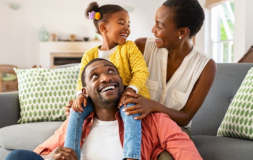 A man with a child sitting on his shoulders. A woman is sitting on a sofa behind the man. The child and woman are smiling at each other