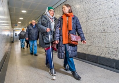 Two women walking. One is using a stick and is being helped by the other